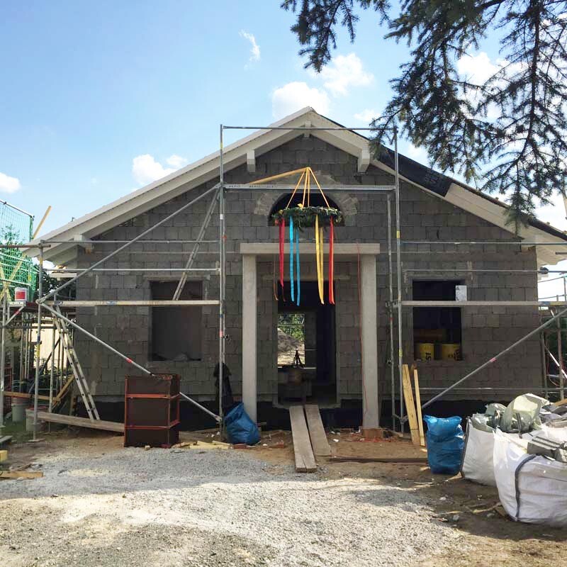 Rohbau eines Hauses mit Gerüst; bunter Kranz mit Bändern über dem Eingang.