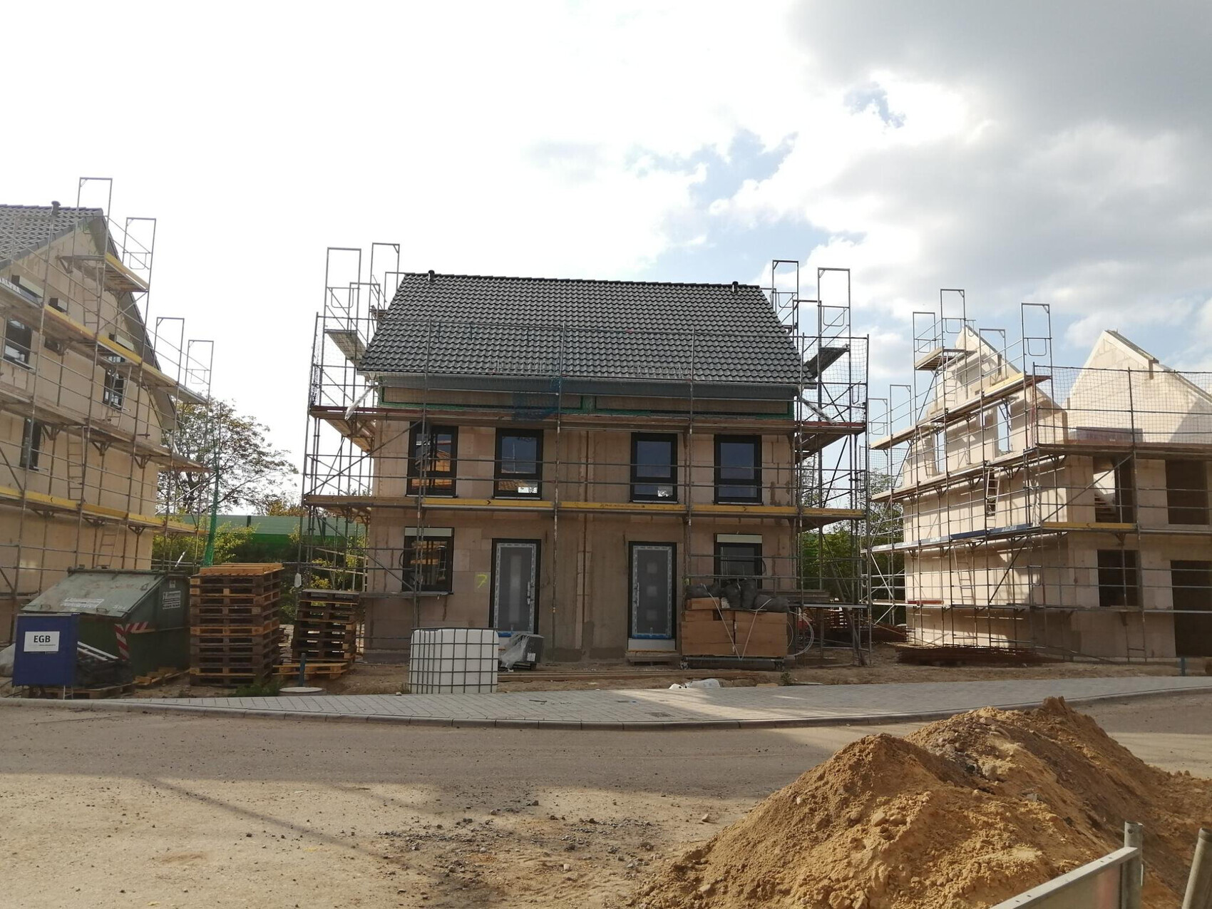 Drei Rohbauhäuser mit Gerüst auf einer Baustelle; Paletten, Erdhaufen im Vordergrund.