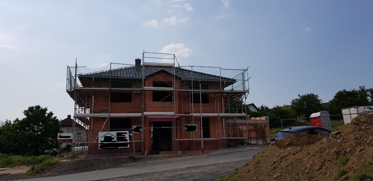 Mehrstöckiges Haus im Rohbau mit Gerüst, Baustellenzaun und Schutt am Grundstück.