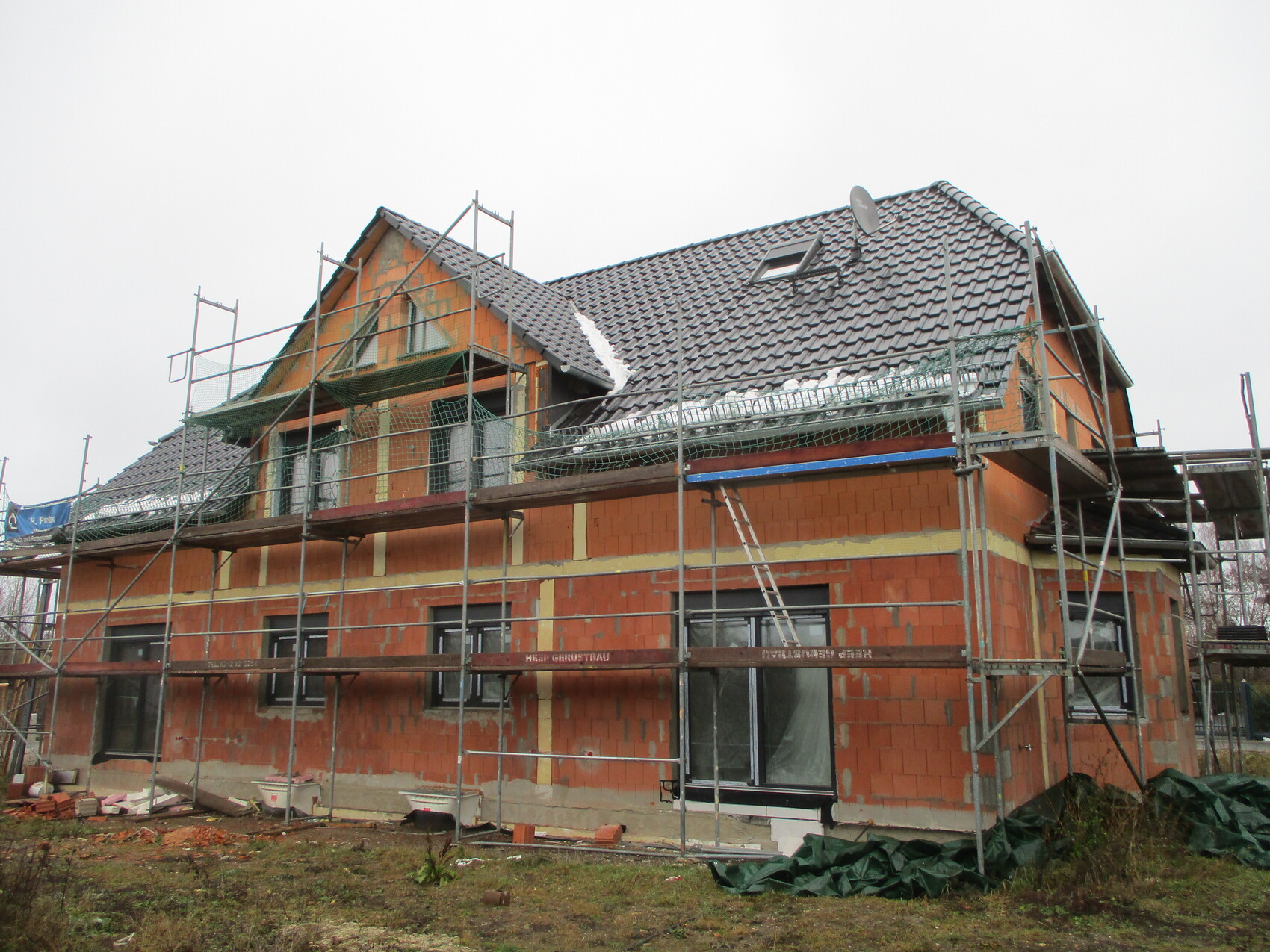 Rohbau eines Hauses mit Gerüst, orange Backstein, Ziegeldach und Bauutensilien auf dem Gelände.