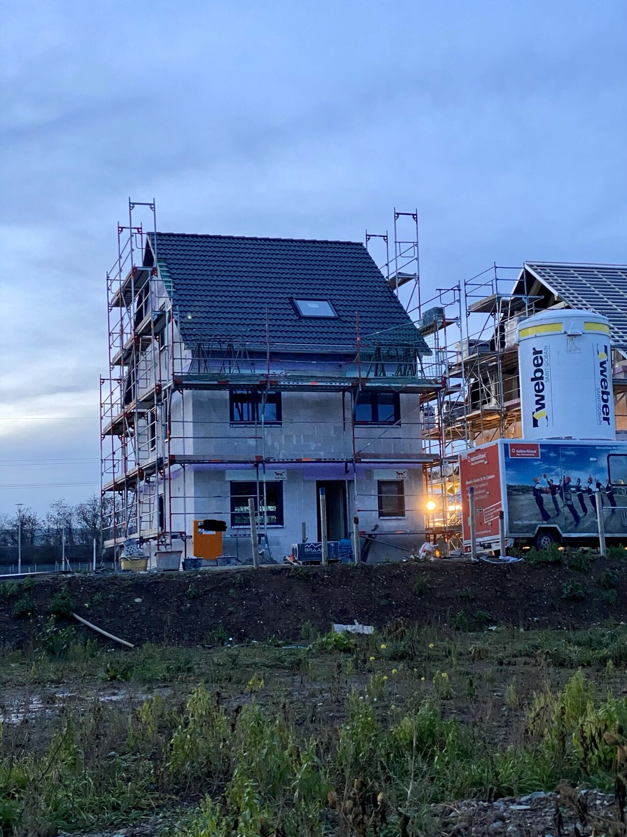 Zweistöckiges Haus im Bau mit Gerüst, dunklem Ziegeldach, Baucontainer.