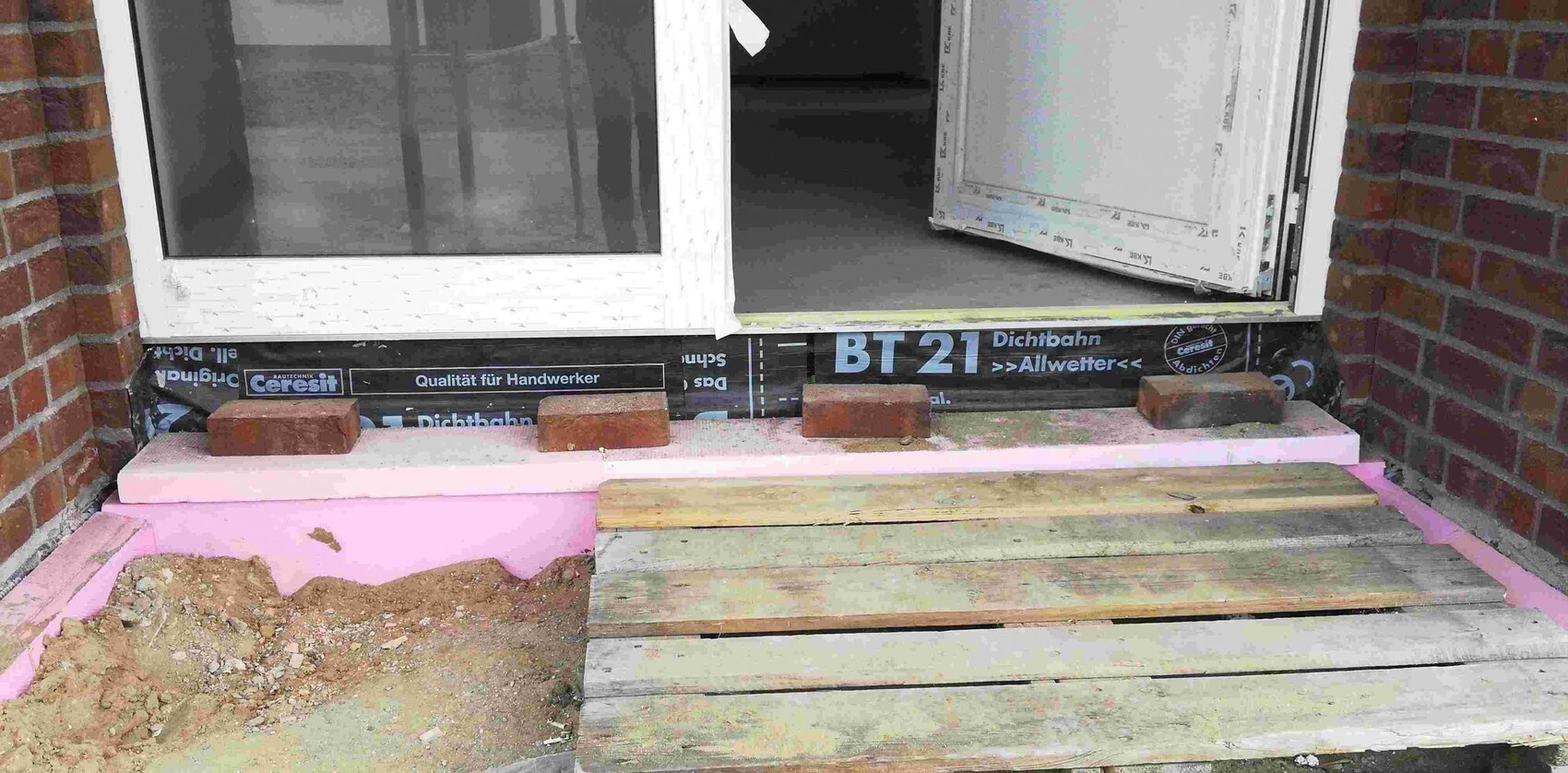 Baustelle am Haus: offener Türrahmen und Fenster mit Holzbrettern, Ziegeln und Sandboden am Eingang