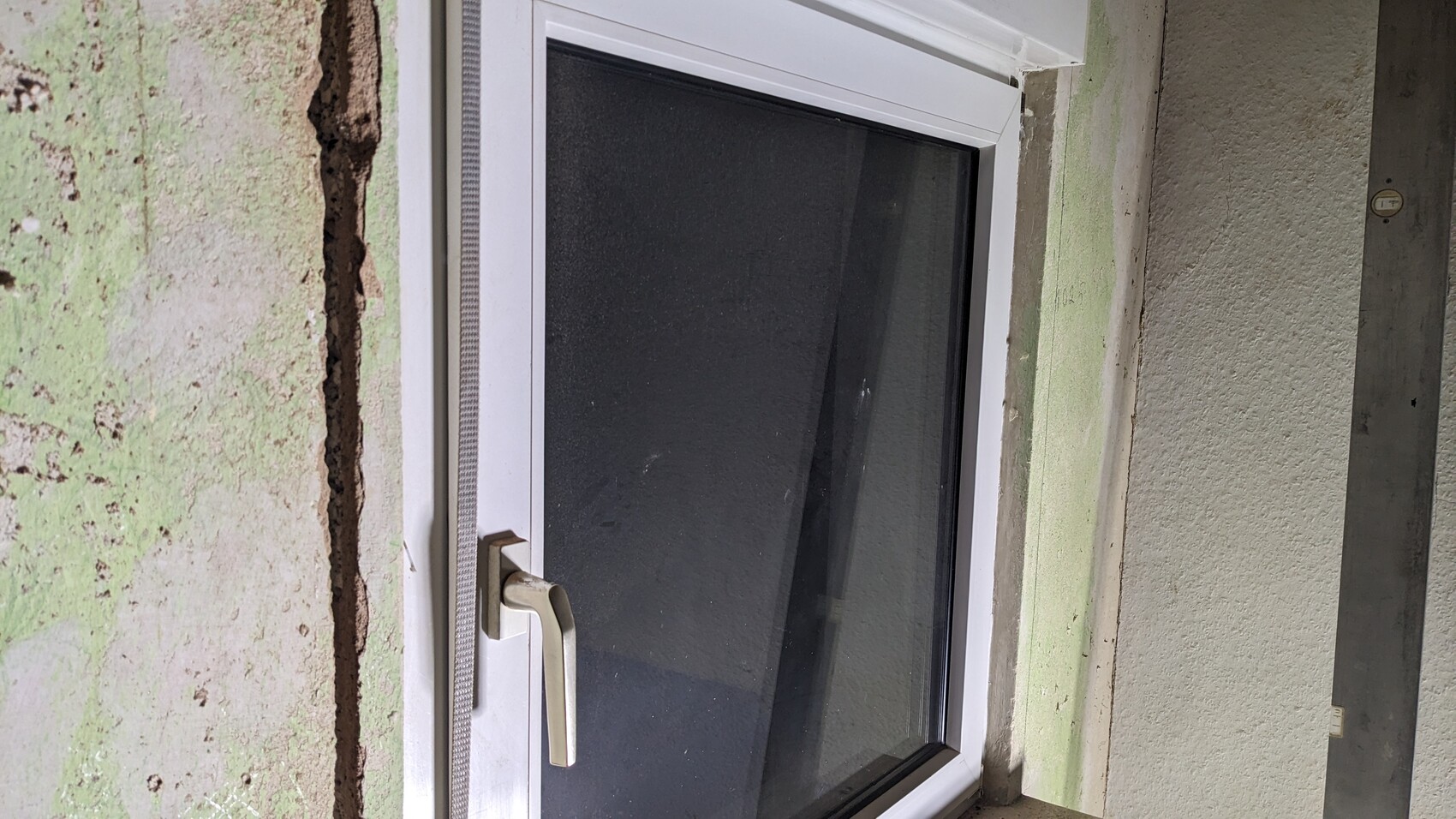 Weißer Fensterrahmen mit Griff vor grünlich verputzter, rissiger Wand.