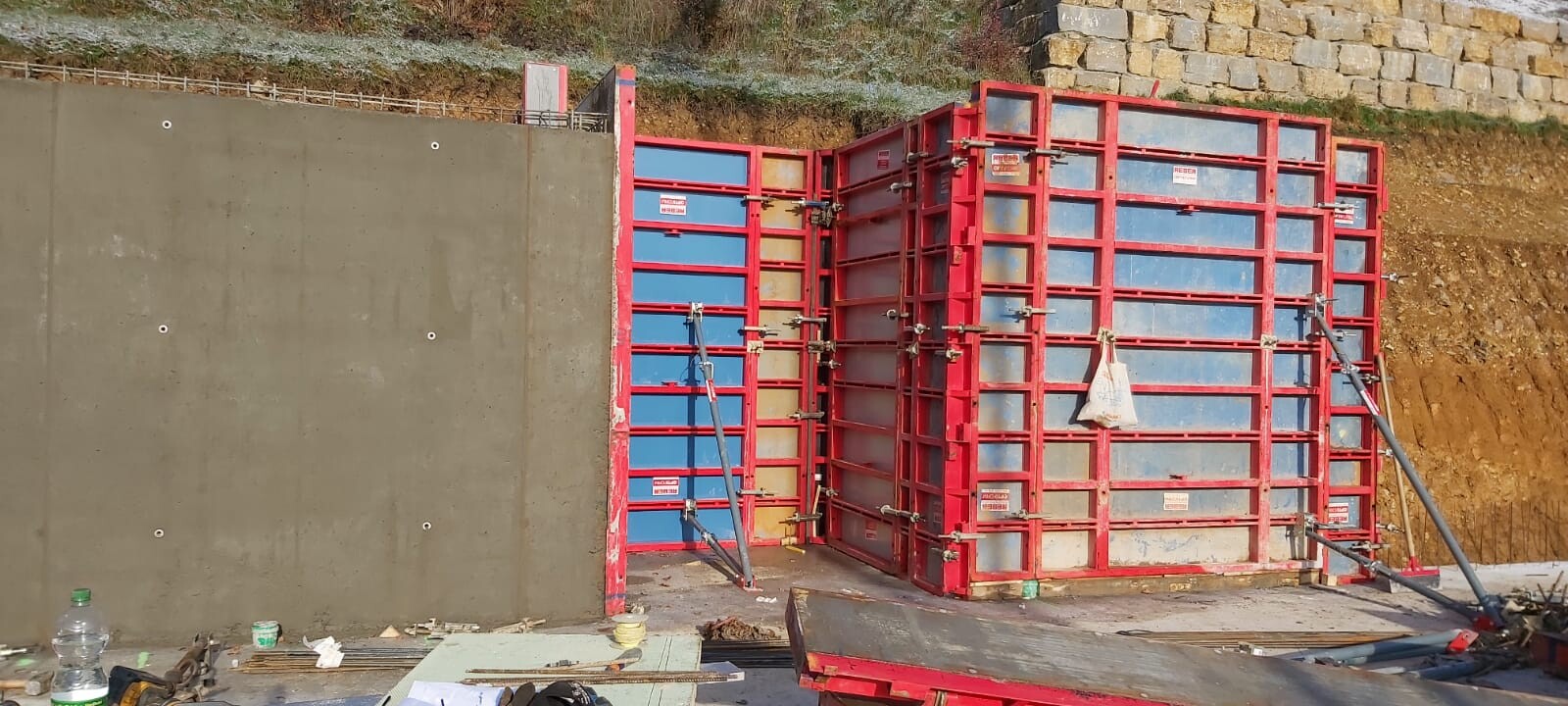 Baustellenschalung mit roten Metallrahmen und blauen Paneele, Gerüstarbeiten und Werkzeuge am Boden.