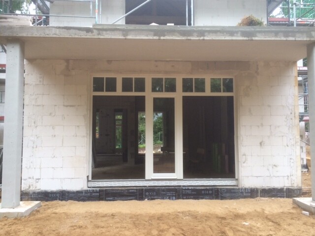 Unfertiges Haus mit großen Glas-Türen, weißen Rahmen, Baugerüst und Erdreich davor.