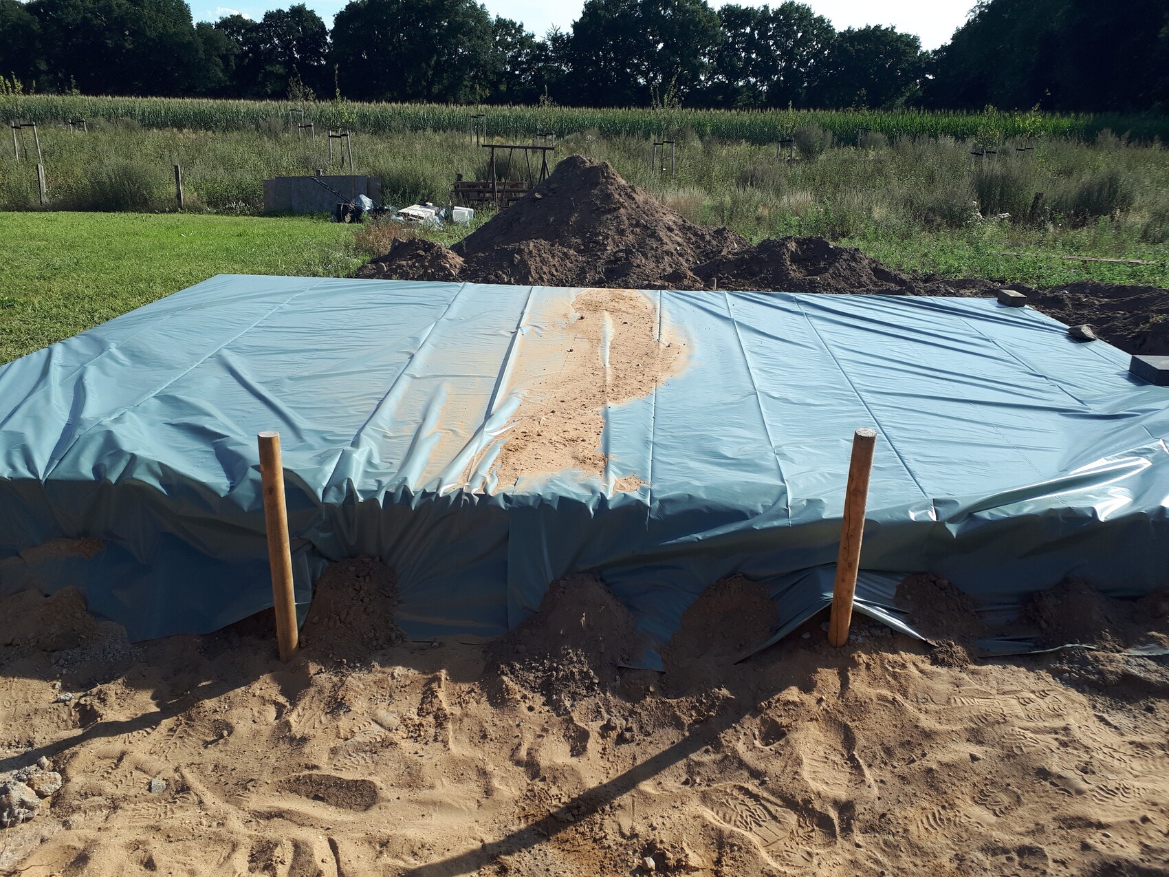 Fundament-Grube mit blauer Abdeckung, Sandhügel und Holzpfosten vorn; Bauplatz im Freien.