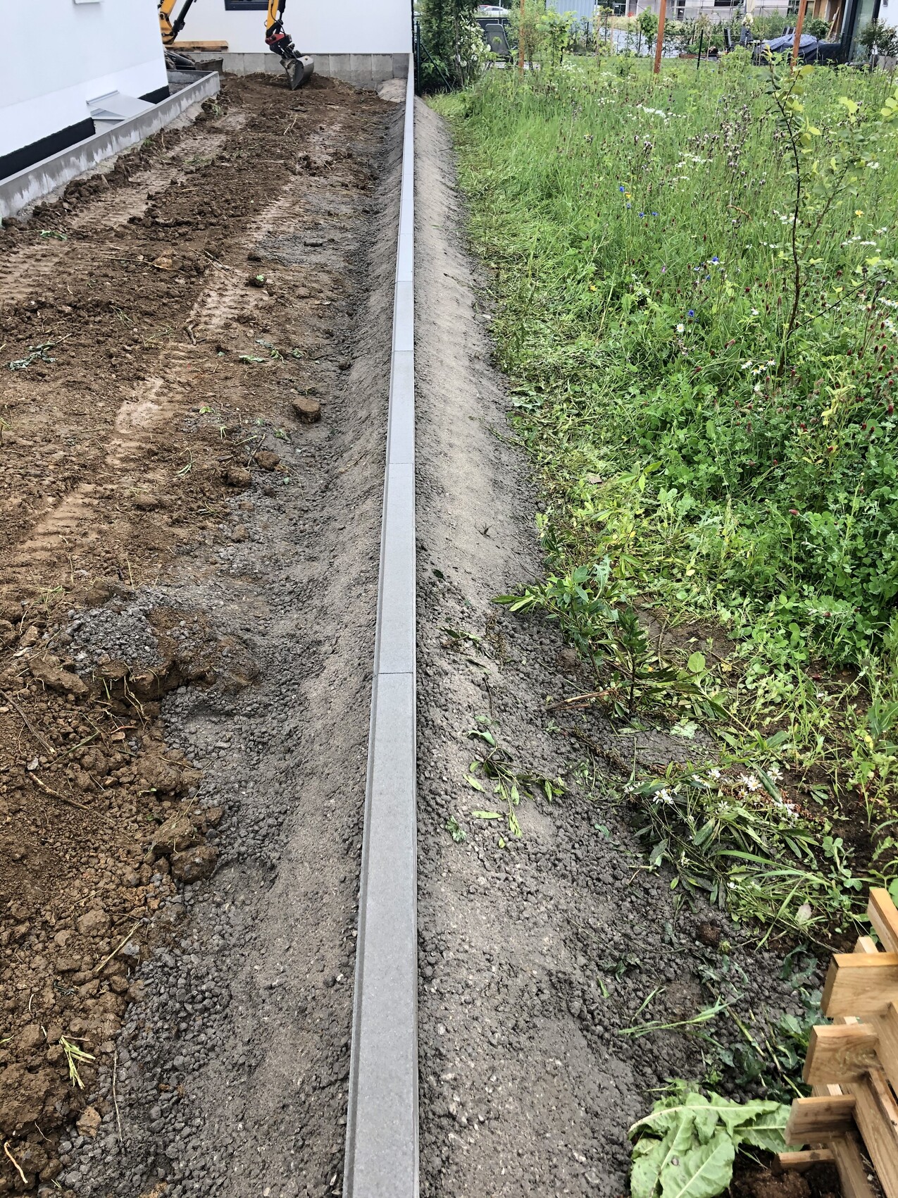 Langgezogener Betonrand trennt Baugrube (links) von Grasfläche (rechts), Baugerät sichtbar.