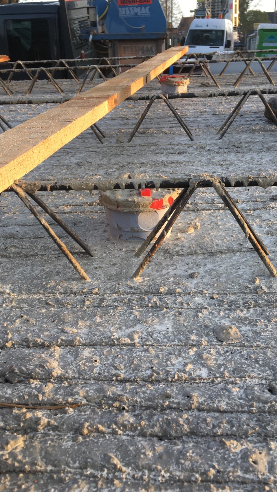 Diagonal liegender Holzbalken auf Stahlrahmen über Betonbaustelle; Hintergrund mit Baugeräten.