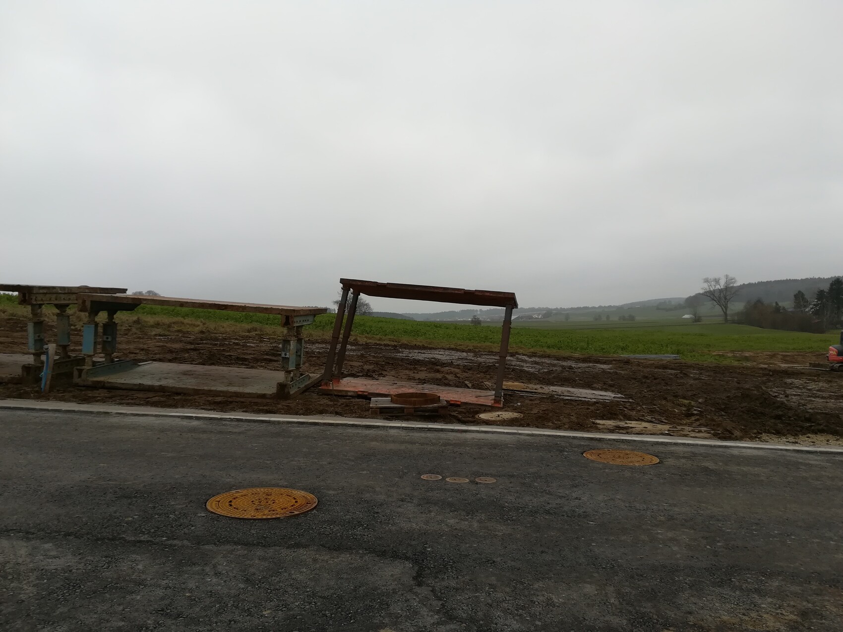 Baustelle am Straßenrand: Holz- und Metallrahmen, schlammiger Boden, Felder im Hintergrund.