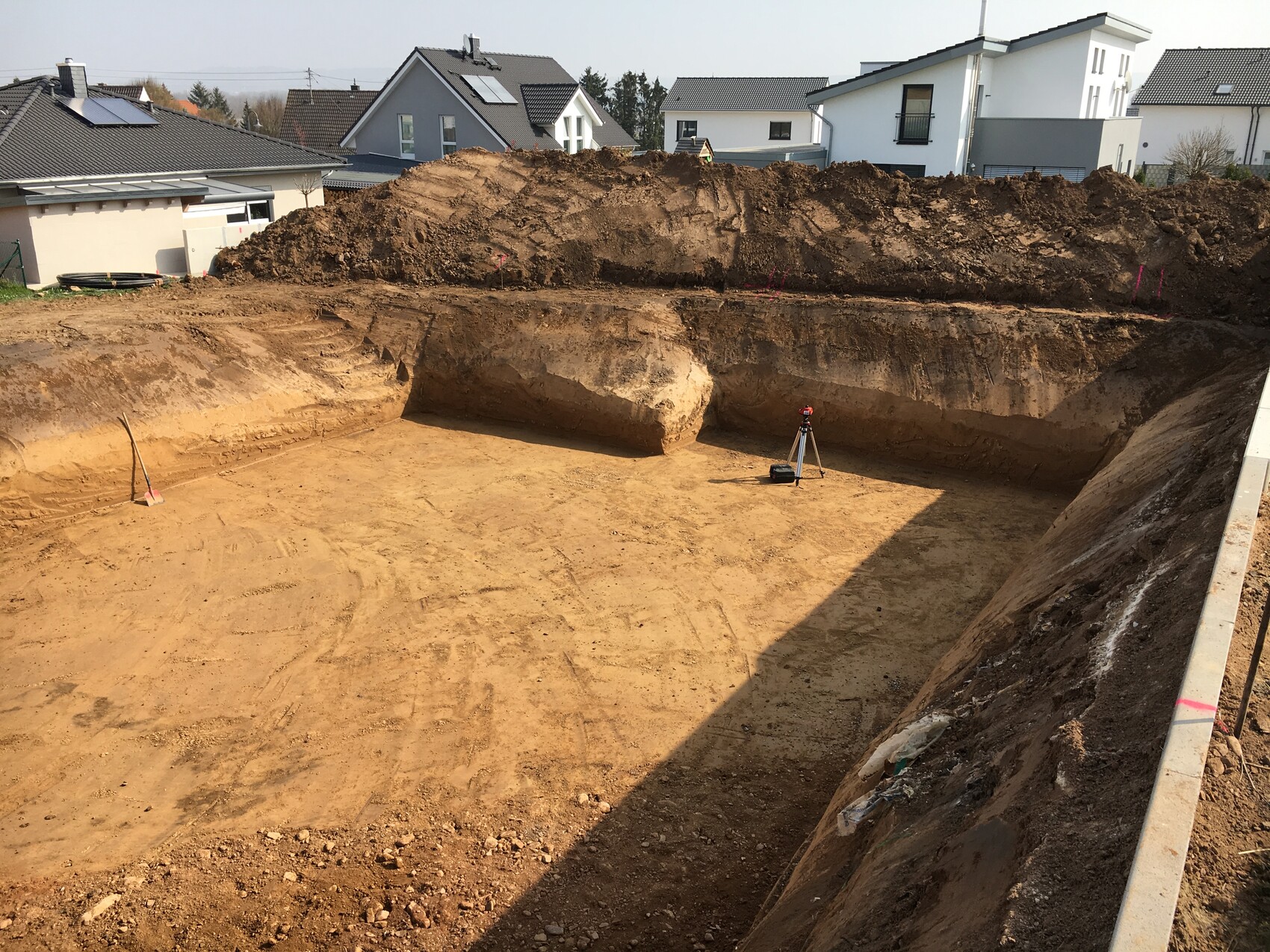 Große Baugrube in Wohngebiet; Erdarbeiten, Messgerät auf Stativ, Hintergrundhäuser.