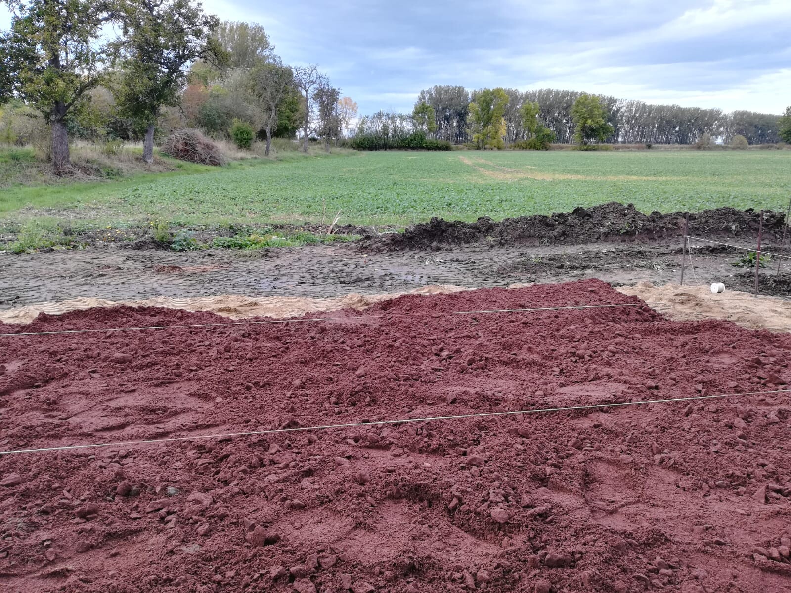 Roter Erdhügel und Graben auf einer Baustelle im ländlichen Feld, Bäume im Hintergrund.