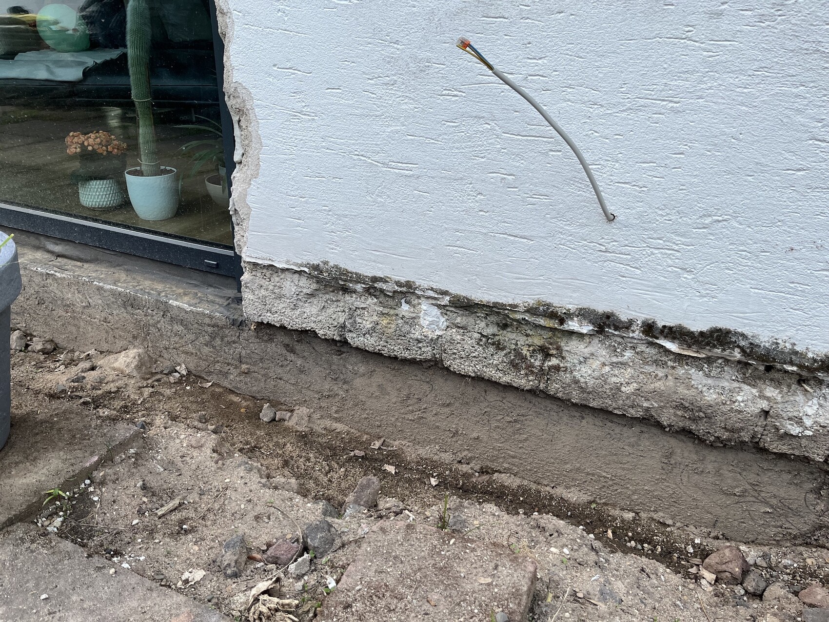 Außenwand mit freigelegtem Fundament, Erdreich darunter, Kabel aus Wand, Glastür links.
