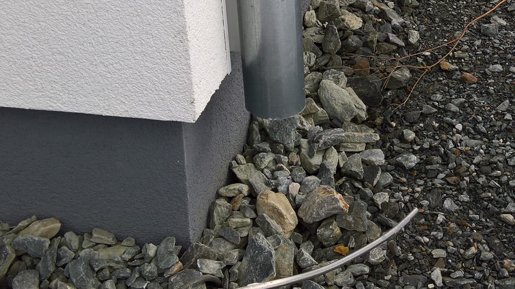 Außenwand-Fundament mit dunklem Sockel, umliegende Kieselsteine und Abflussrohr