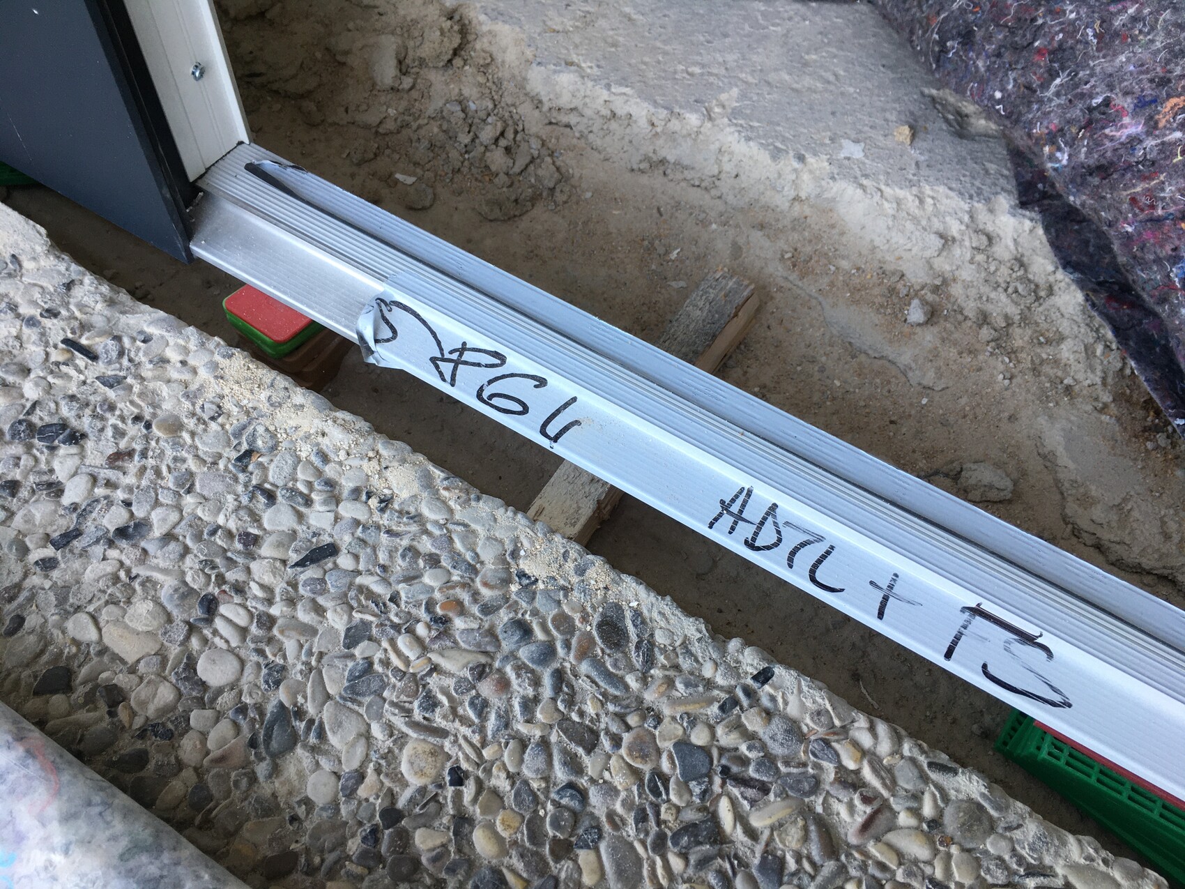 Aluminium-Schiene als Türschwelle über einer Baugrube; Kiesboden im Vordergrund, Graffiti auf dem Metall.