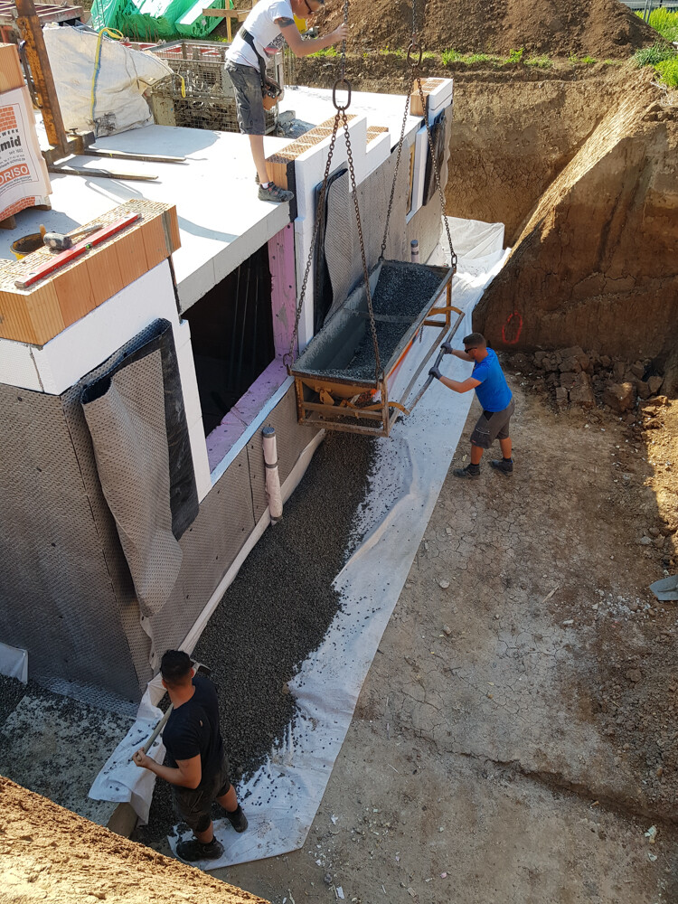 Baustelle: Betonfundament wird gegossen, Kippmulde hängt an Ketten, Arbeiter arbeiten.