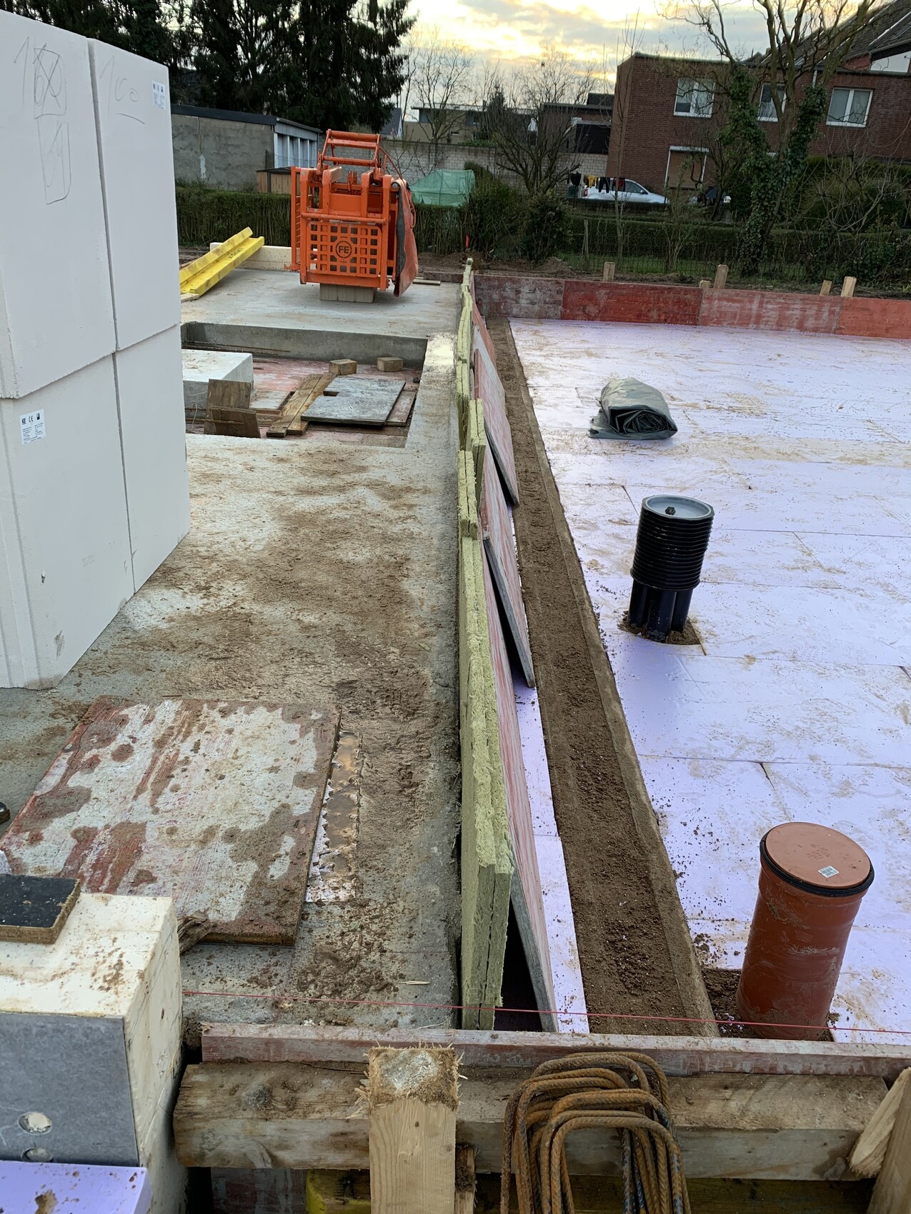 Baustelle: Fundamenttrasse mit pinker Abdichtbahn, orange Baumaschine, Holzplatten.