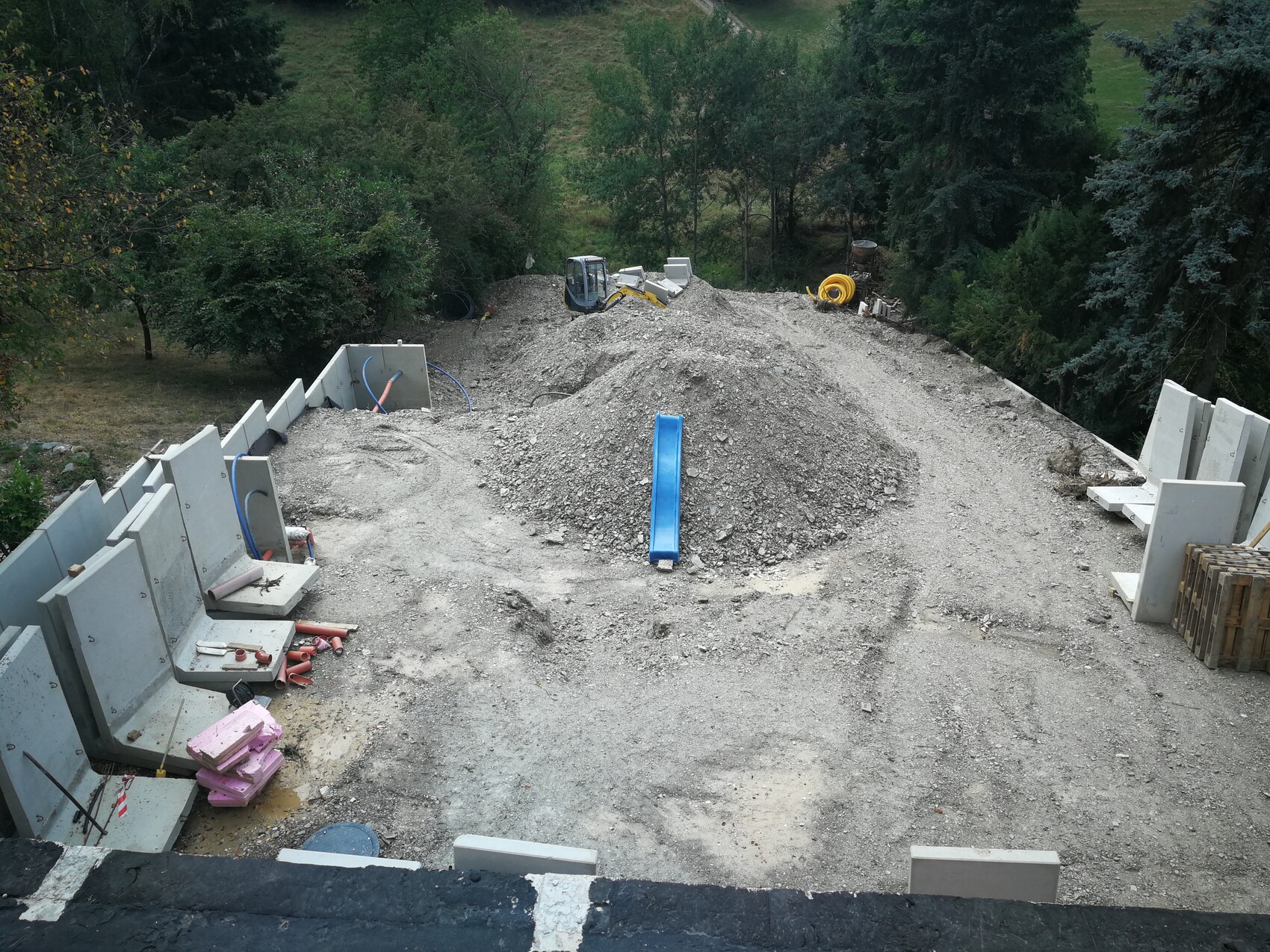 Baustelle: Kieshügel, blaue Rutsche, Betonwände, Baumaschinen im Hintergrund.