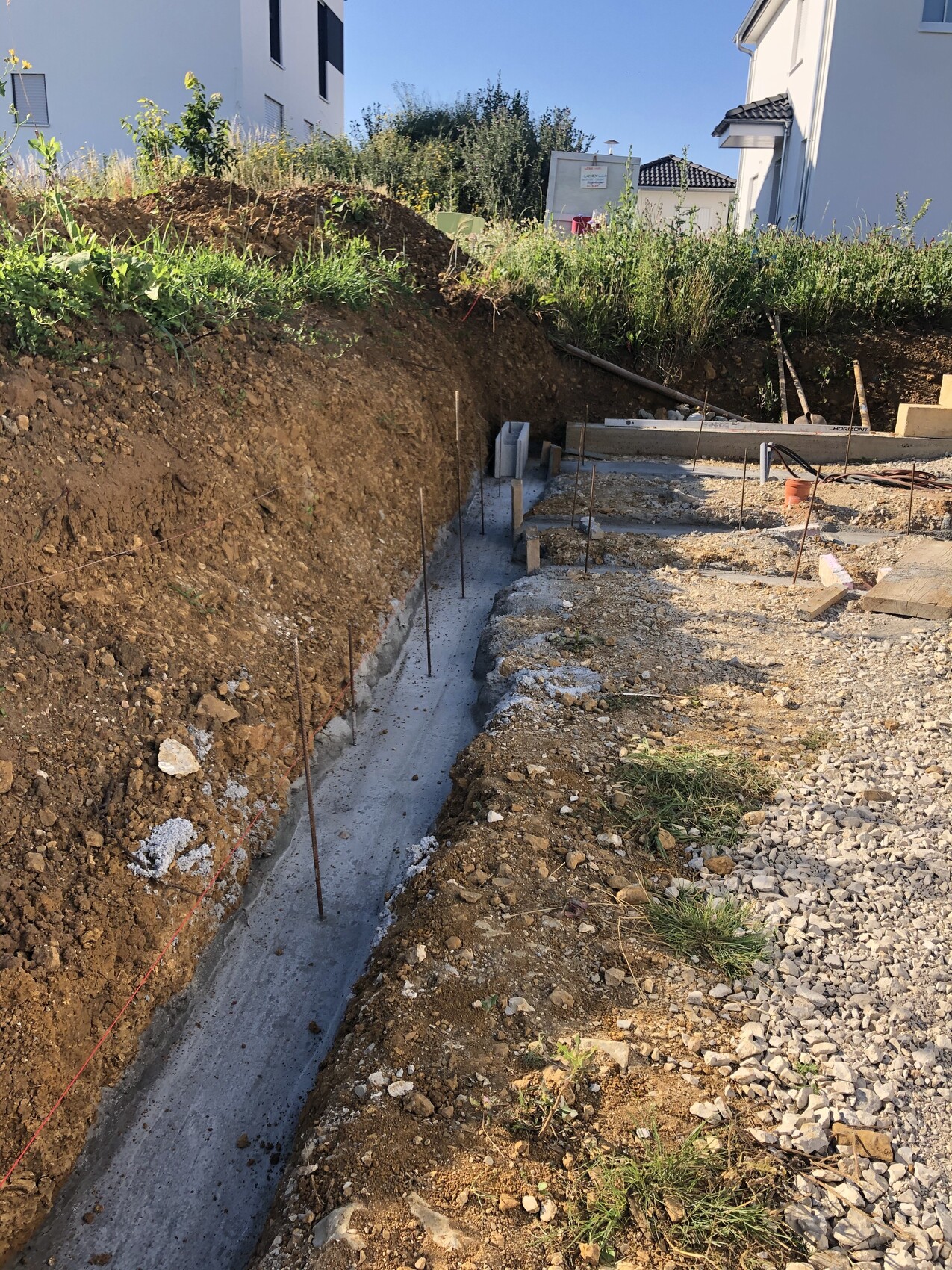 Baustelle mit tiefem Graben, frisches Betonfundament mit Bewehrungsstangen, Kies und Erde.