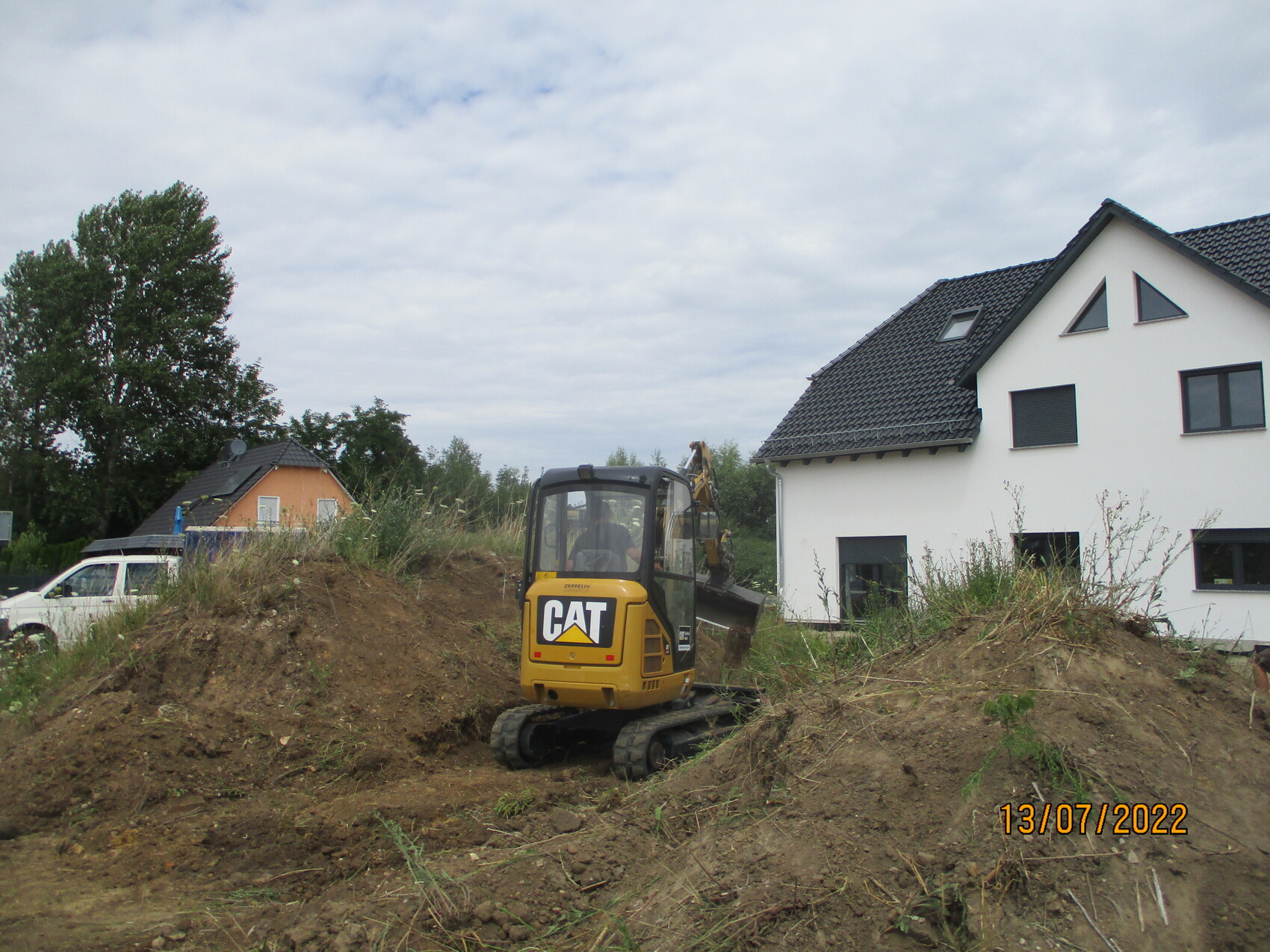 Kleine Baustelle: gelber CAT-Bagger auf Erdhügel neben weißem Einfamilienhaus, Datum 13/07/2022.
