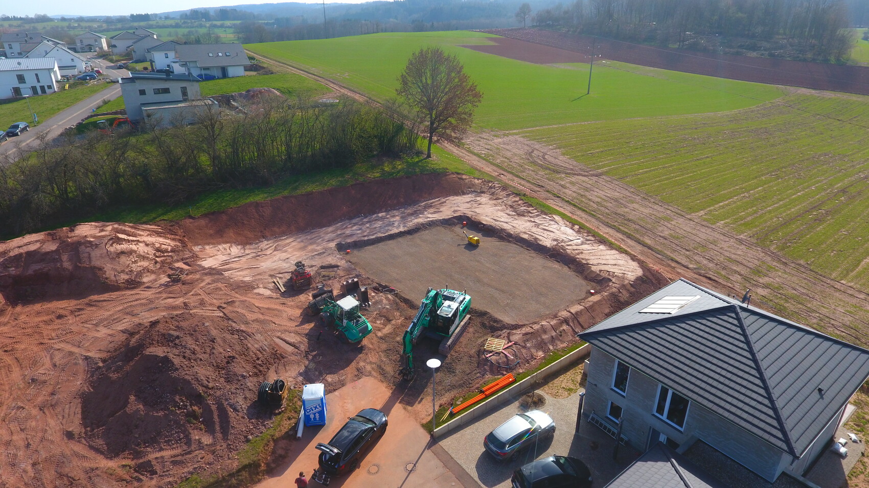 Großes Baugelände mit Erdarbeiten, Baumaschinen Bagger Radlader neben Wohnhaus, Felder im Hintergrund.