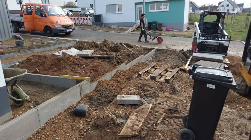 Baustelle: Erde, Betonplatten, Paletten; Bauarbeiter schiebt Schubkarre, LKW im Hintergrund