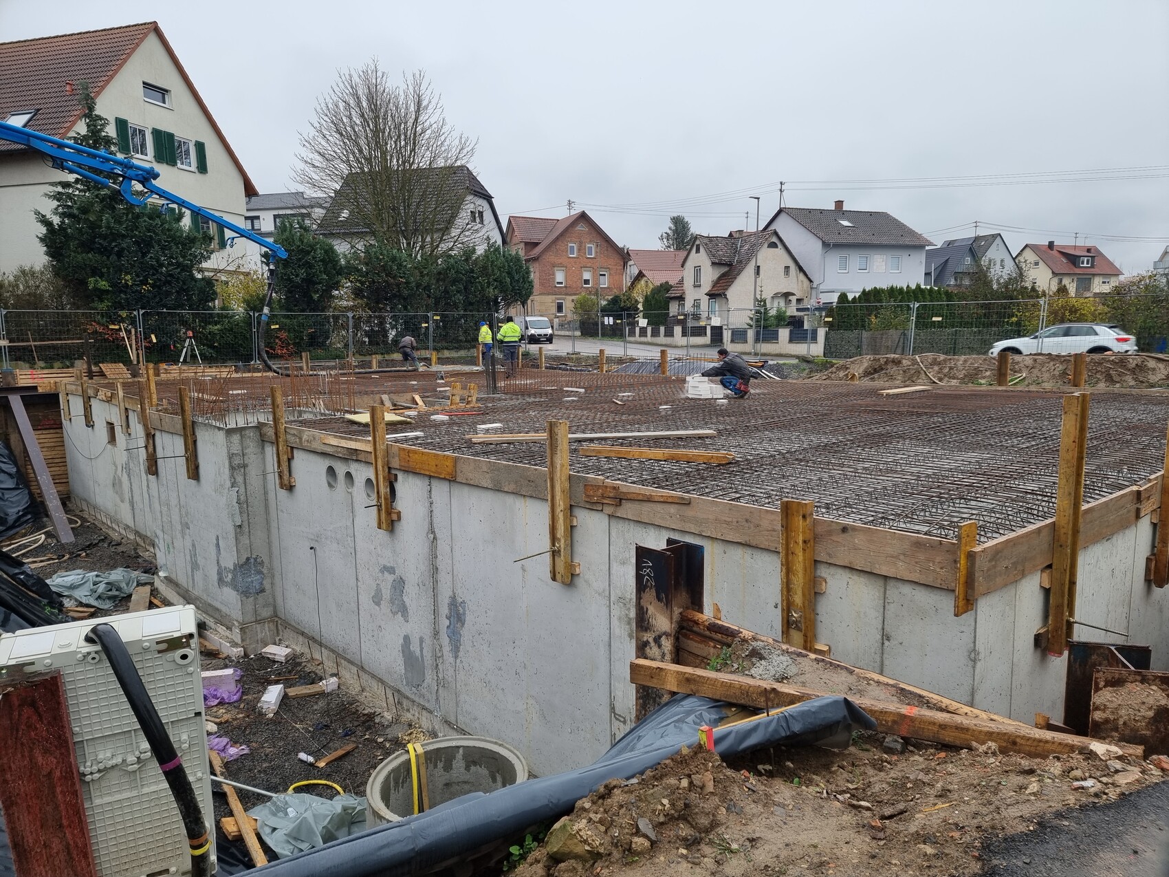 Baustelle mit Fundament, Stahlbewehrung, Holzformen und Bauarbeitern im Hintergrund.