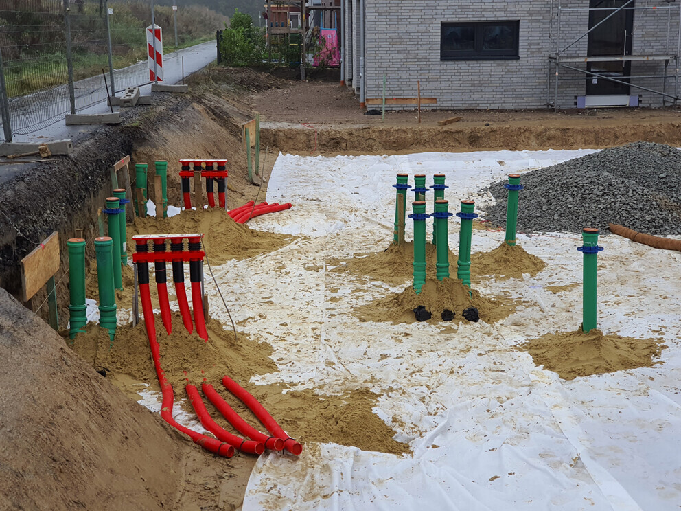 Baugrube mit grünen und roten PVC-Rohren, Sandschichten und Erdarbeiten, Gebäude im Hintergrund.