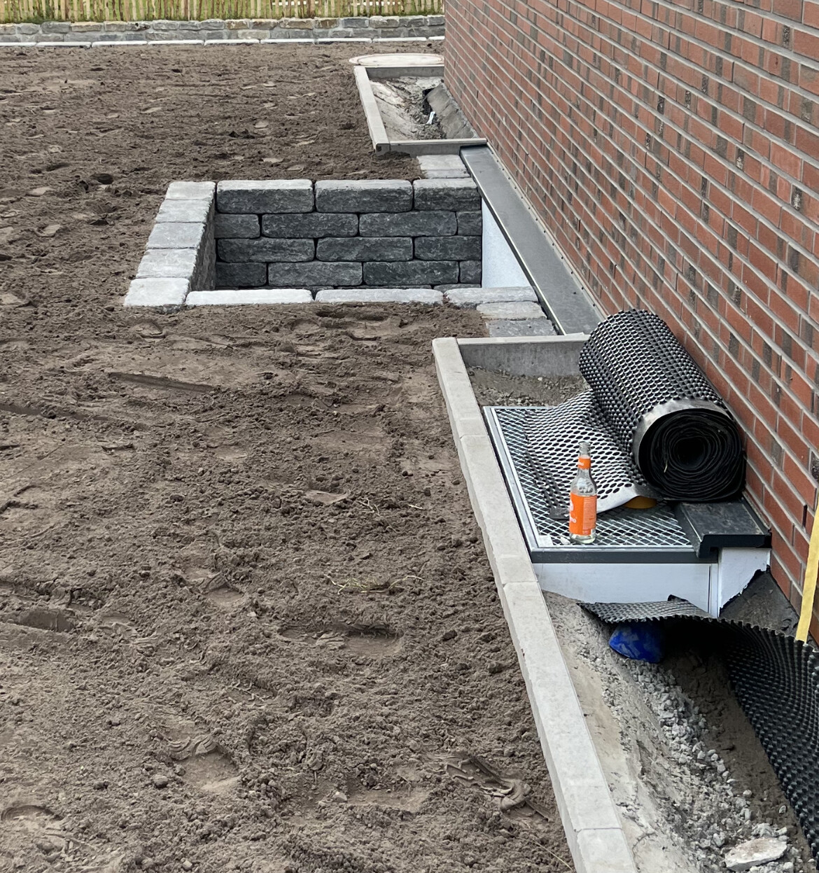Bauplatz am Haus: niedrige Steinmauer, aufgerollte Dachbahn auf Metallgitter, Flasche daneben.