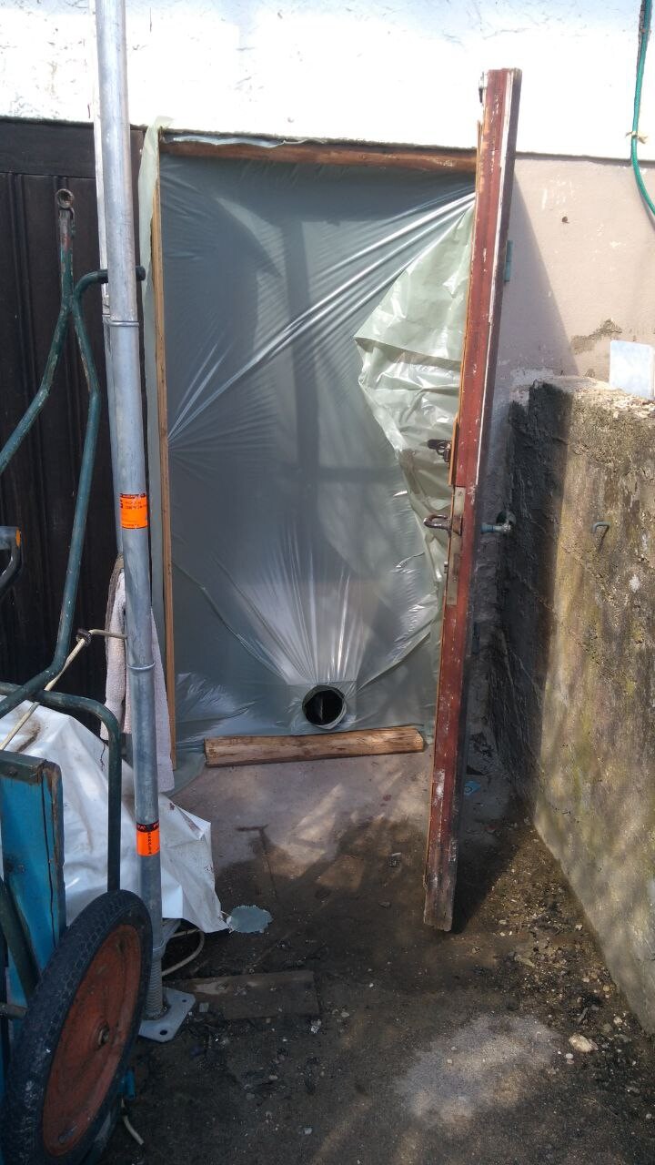 Provisorische Türöffnung hinter Plastikfolie, Holzlatte unten, Schubkarren davor.