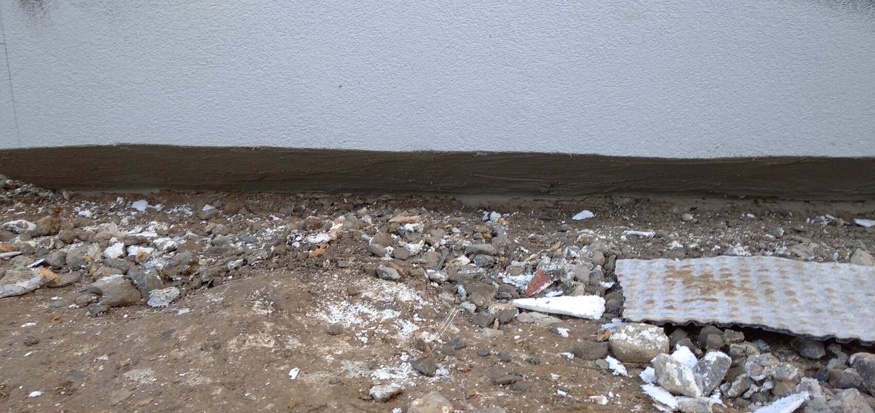 Unterer Fassadenbereich mit braunem Sockel; Kies, Geröll und Baumaterialreste am Boden.