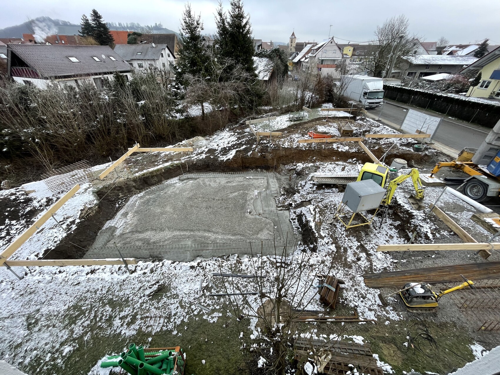 Baustelle mit Fundamentplatte, Holzformen, Bagger und LKW, Schnee bedeckt, Wohngebiet.