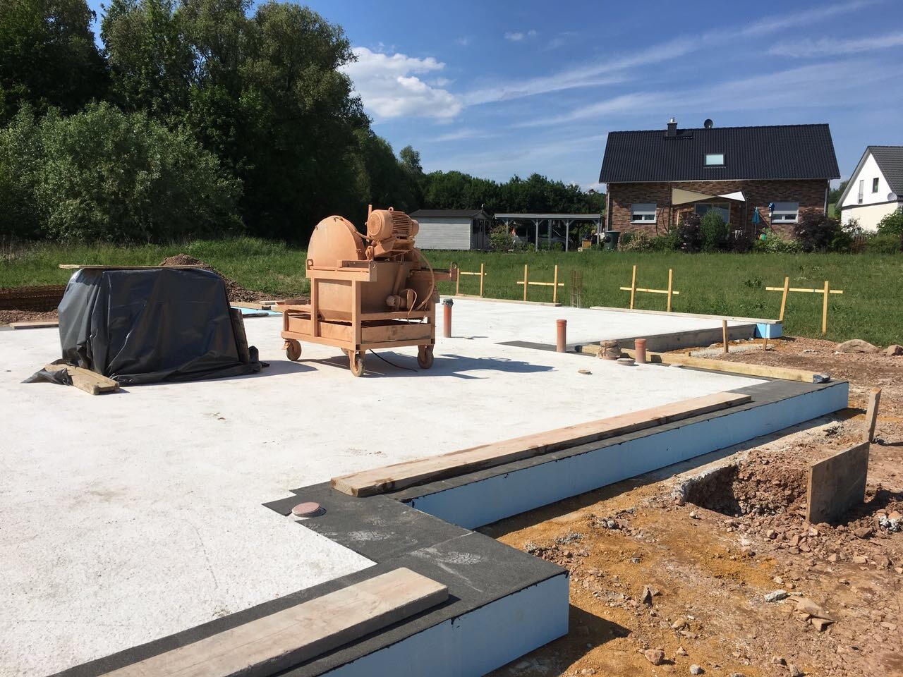 Baustelle mit Betonfundament, Betonmischer, Holzlatten und Pfosten vor Häusern im Hintergrund