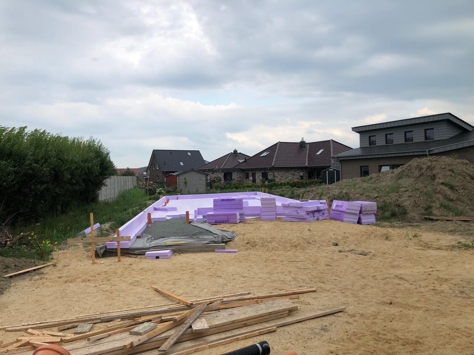 Baustelle mit Fundament und gestapelten lila Dämmplatten, Holzbretter, Häuser im Hintergrund.