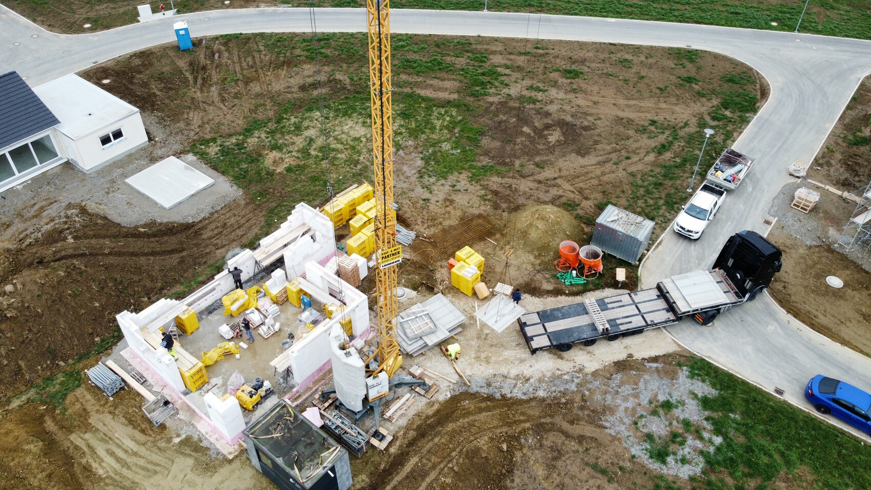 Vogelperspektive: Gelber Baukran ueber halbfertiger Baustelle mit Betonwaenden und Materialstapeln