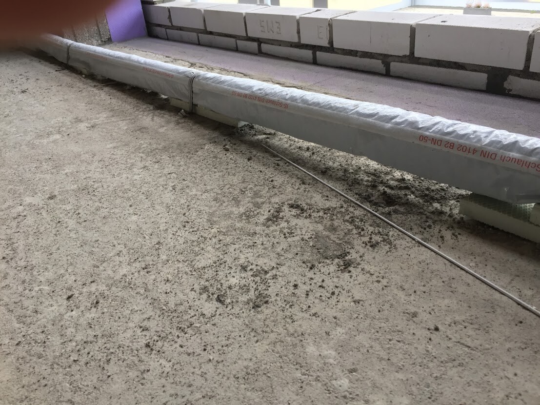 Isoliertes Rohr liegt auf Betonboden, Bauzubehör daneben; im Hintergrund stapeln sich Ziegelsteine.