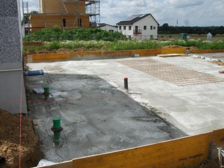 Baustelle mit betonierter Fundamentplatte, Rohrleitungen und Holzformen; Gebäude im Hintergrund.