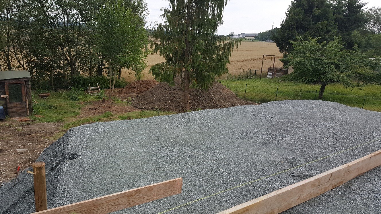 Baustelle mit Kiesfundament, Holzformen und Absperrung, umliegende Bäume und Felder.