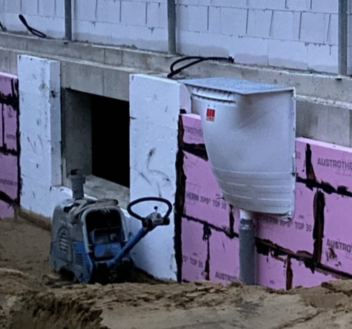 Baustelle: Fundamentwand mit pinken Dämmplatten, Lüftungsschacht, und Vibrationsplatte am Boden