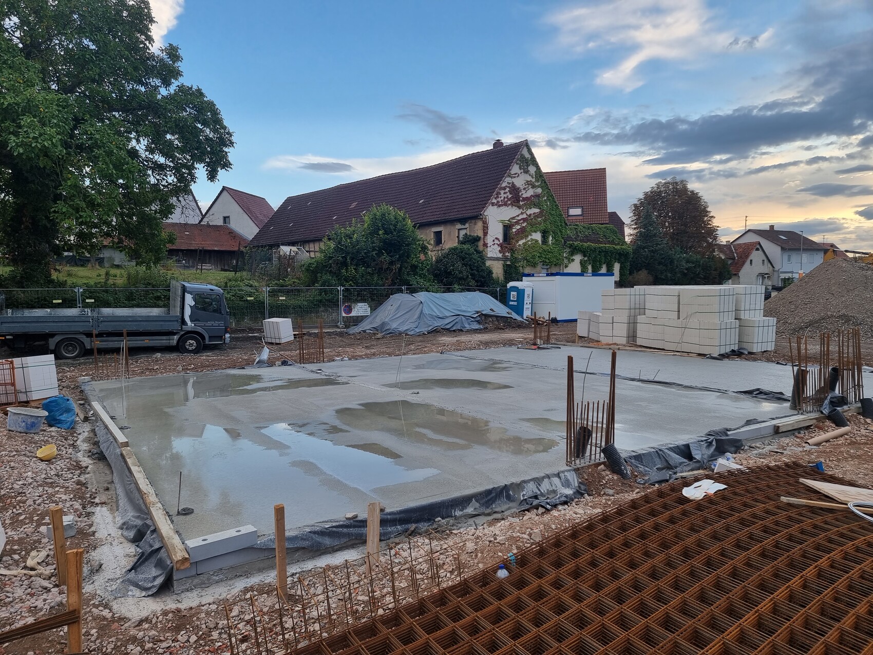 Baustelle mit frisch gegossener Fundamentplatte, Bewehrung, Baumaterial, Häuser im Hintergrund.