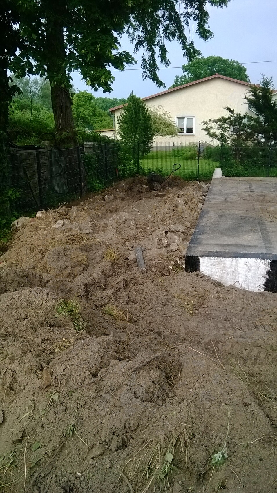 Gartenbaustelle mit Erdaushub, einer Schubkarre und Betonplatte neben Zaun; Haus im Hintergrund.