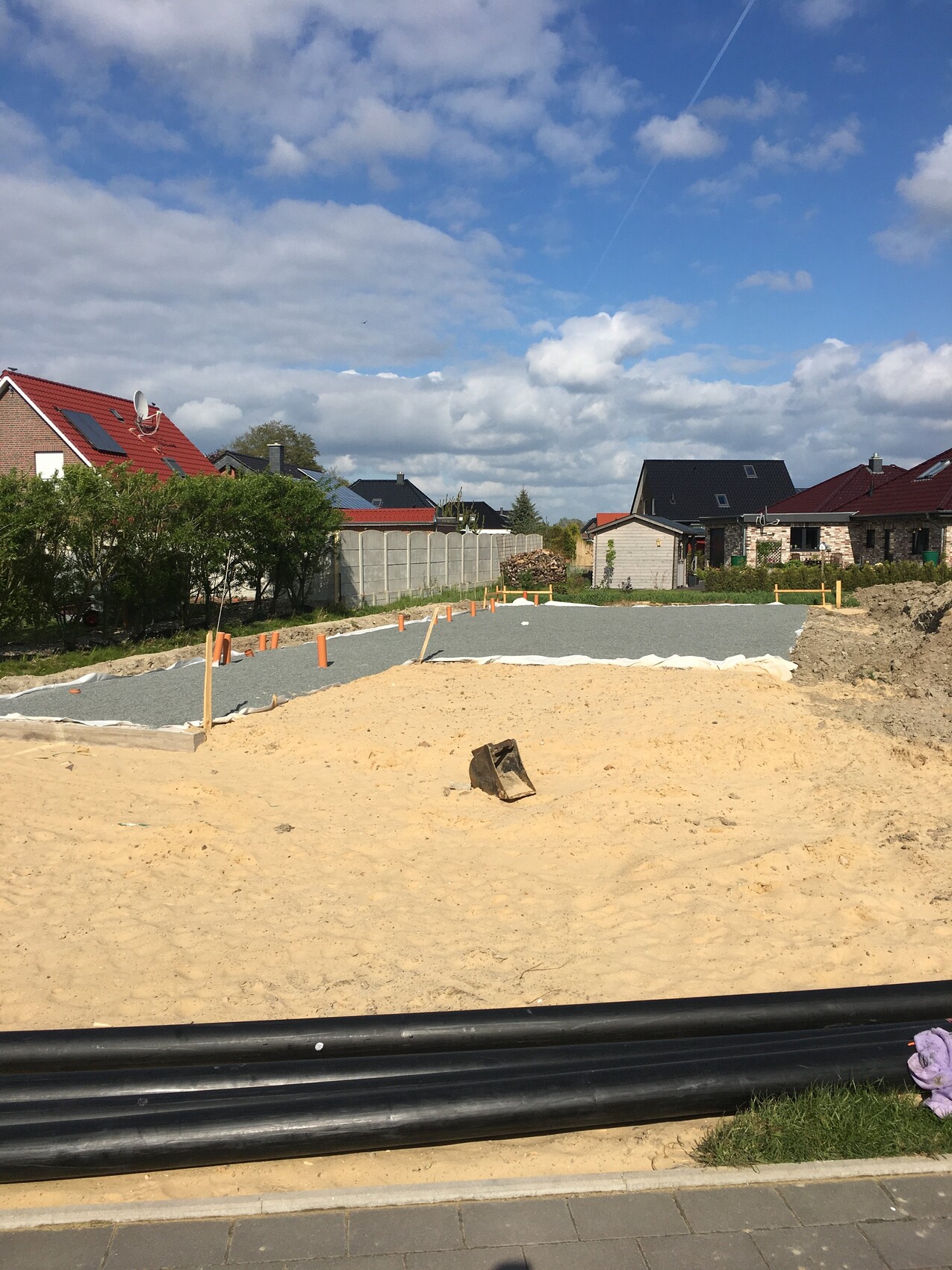 Baustelle in Wohngegend: Kiesweg, Sandhügel, orange Markierungsstangen, Häuser im Hintergrund.