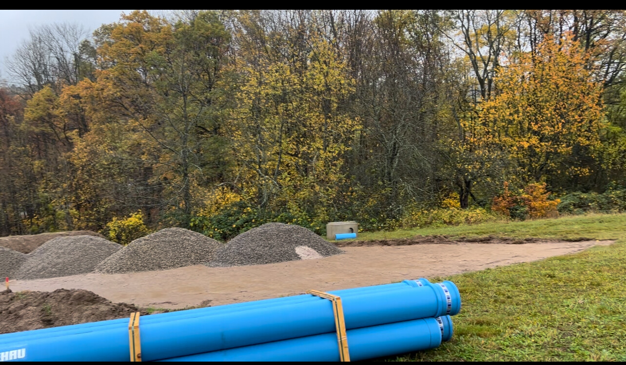 Baustelle mit großen blauen PVC-Rohren, Kieshaufen und Herbstwald im Hintergrund