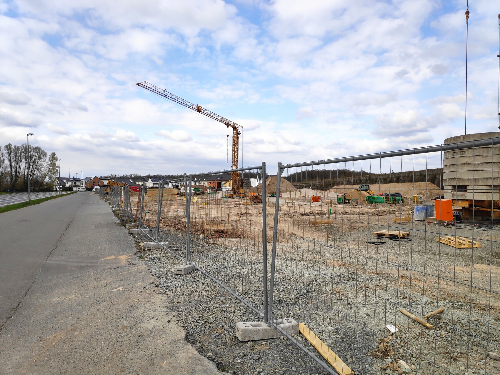 Baustelle mit hohem Kran hinter einem Metallzaun, Kiesboden, Paletten und Baugeräte.