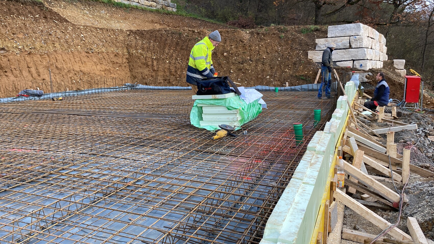 Baustelle mit Bewehrungsstahl, Bauarbeitern, Holzformen und Dämmplatten am Fundament.