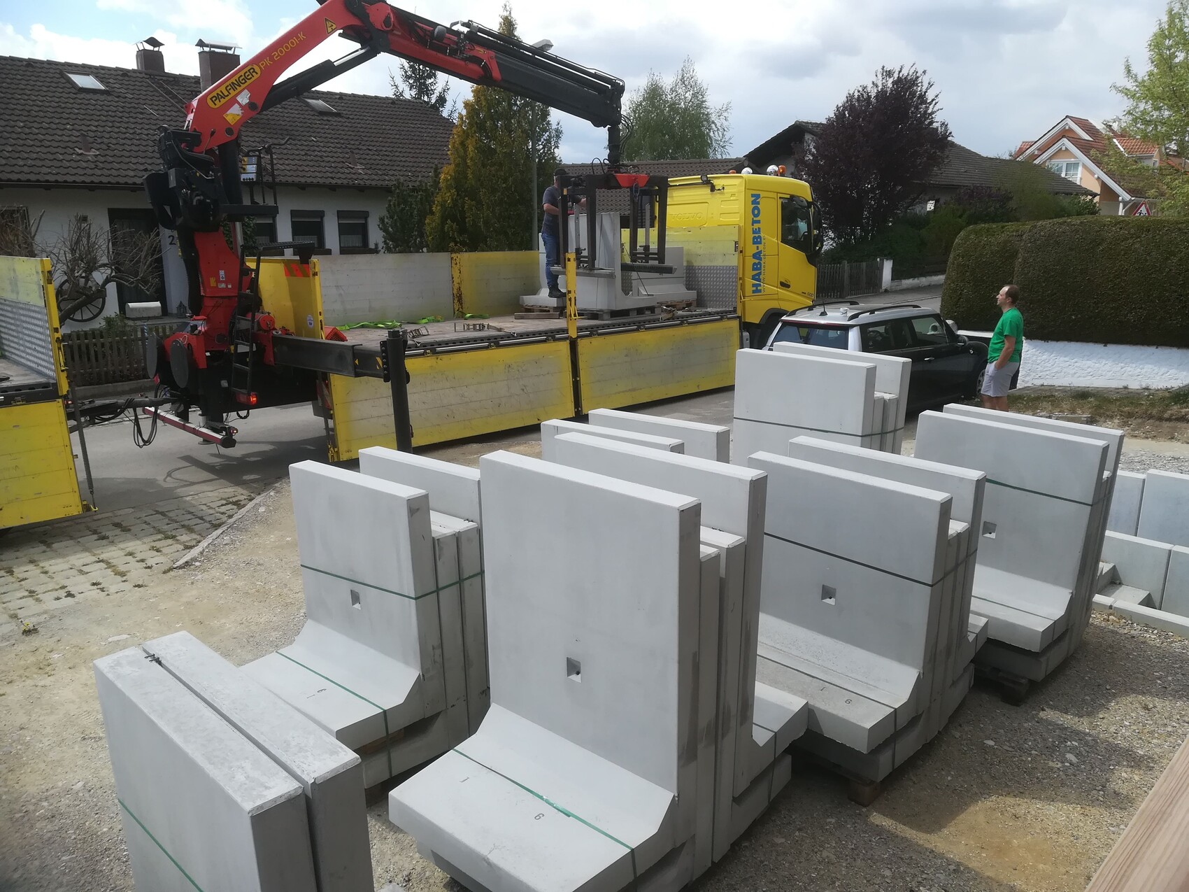 Baukran hebt schwere Betonplatten auf LKW, Baustelle mit Blockstapeln, Mann beobachtet.