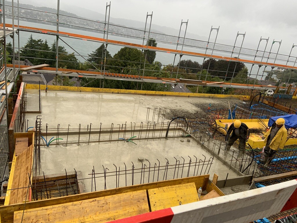 Zwei Bauarbeiter mit Gelbhelm arbeiten auf feuchtem Betonfundament; Stahlbewehrung, Gerüst sichtbar.
