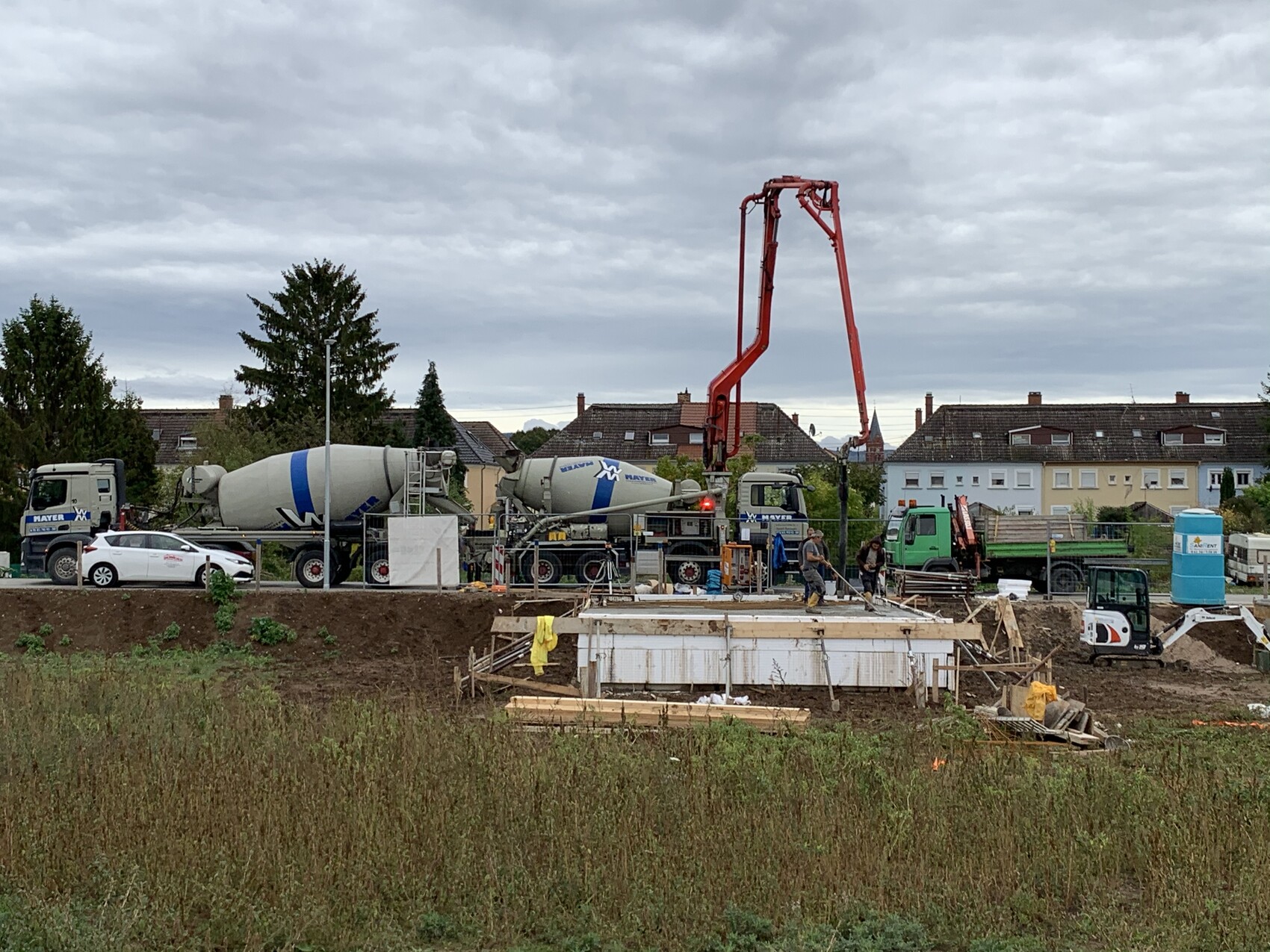 Baugrube mit Betonmischern, rotem Betonpumpenarm und Bauarbeitern beim Fundament