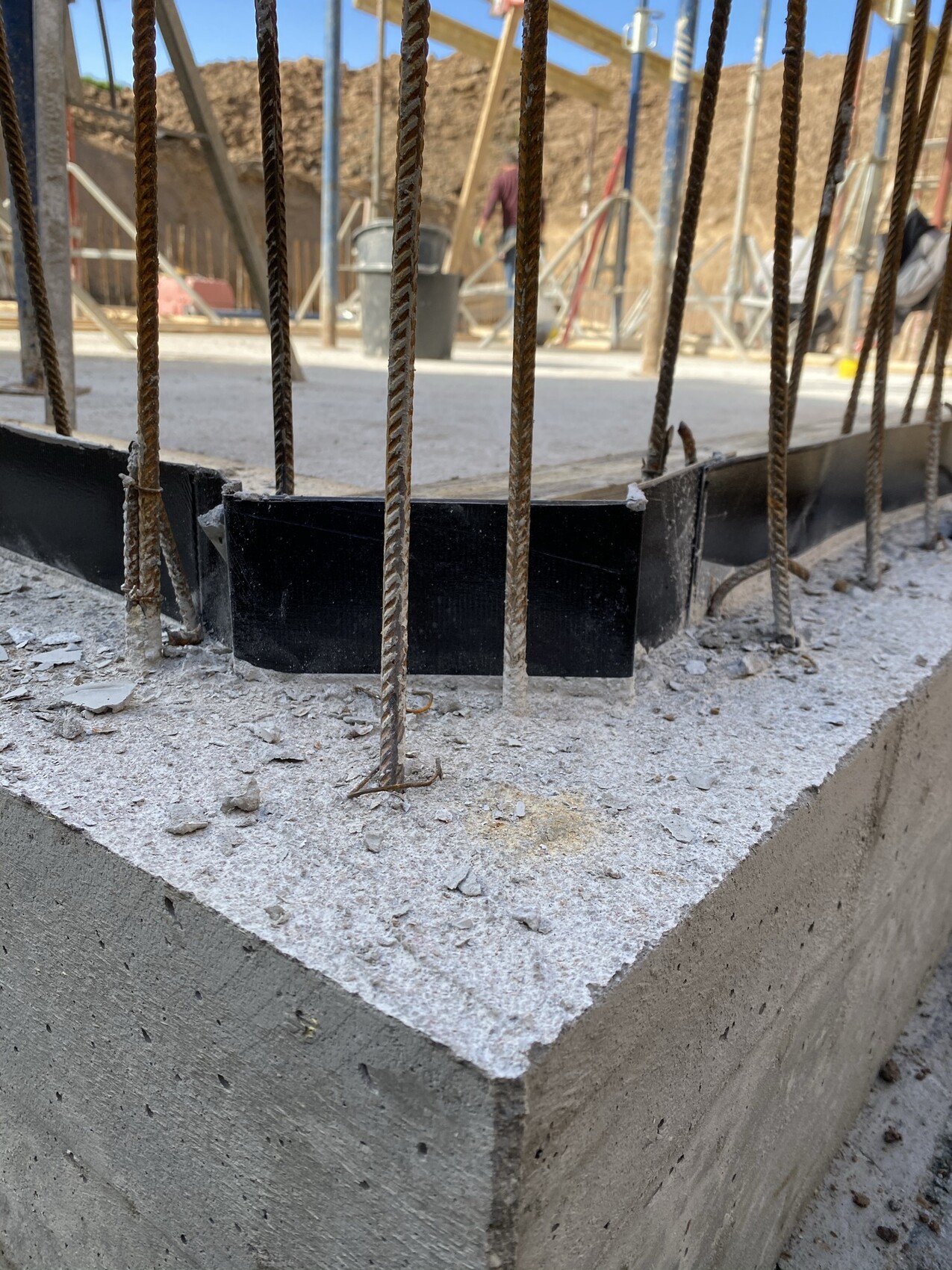 Nahaufnahme einer Betonplatte mit rostigen Bewehrungsstäben; Baugerüst und Arbeiter im Hintergrund.