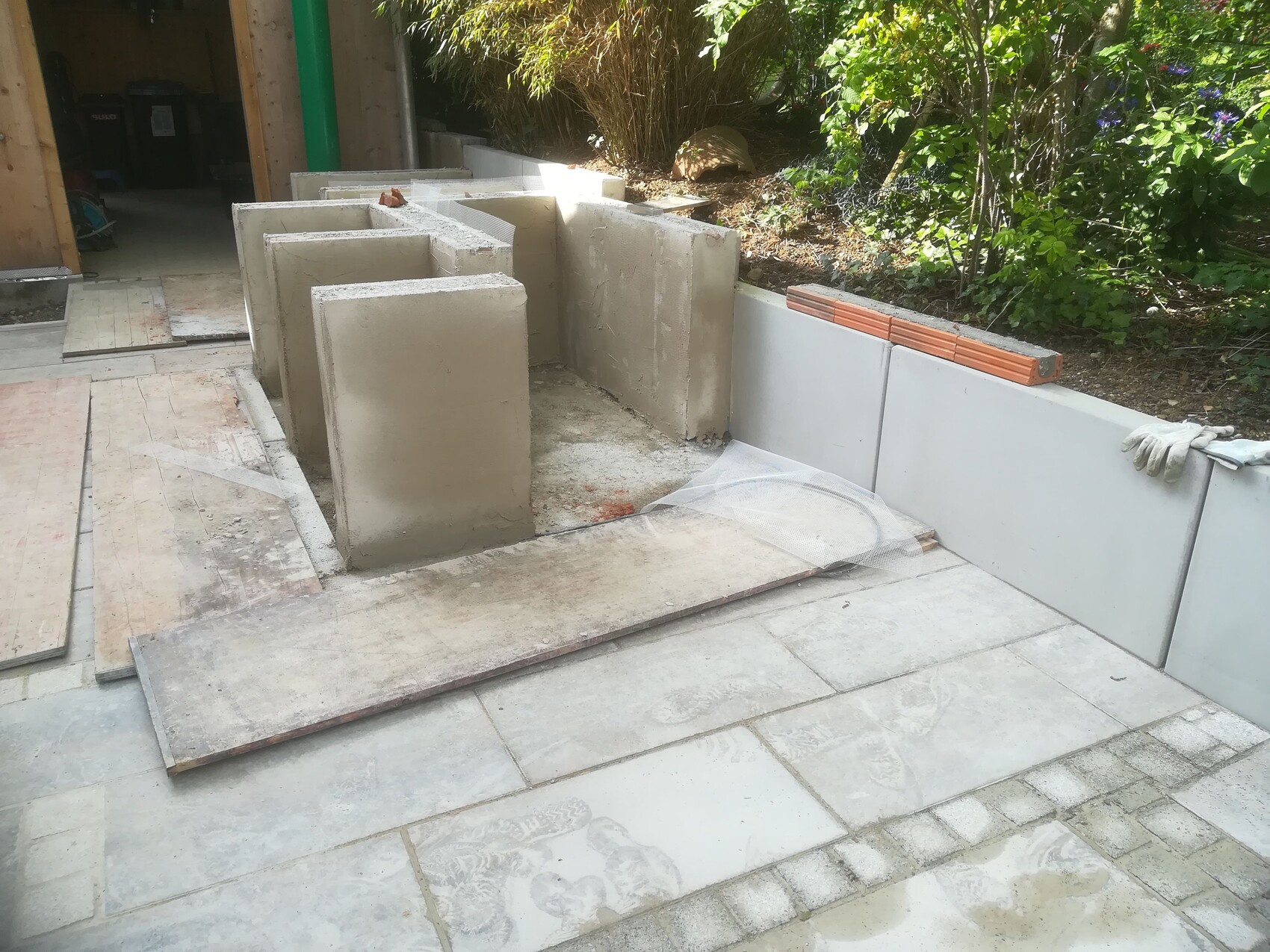 Gartenbaustelle: Betonblöcke formen eine Wand, Holzplatten liegen daneben, Handschuhe auf dem Rand.