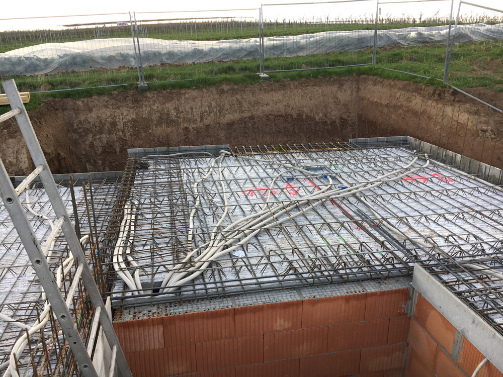 Fundament mit Bewehrungsstahlnetz und Kabelbündeln; Bauzaun rund um die Baustelle.